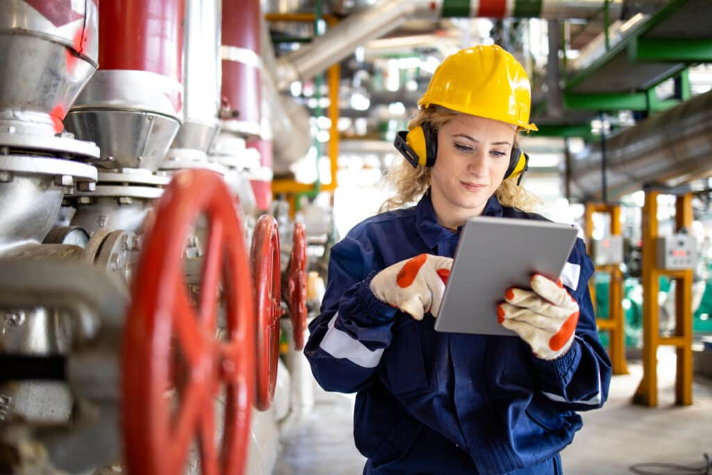 Refinery worker with hardhat and earmuffs checking safety plan on tablet computer for oil and gas production.